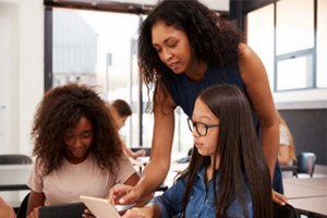 female teacher standing at desk with two female students